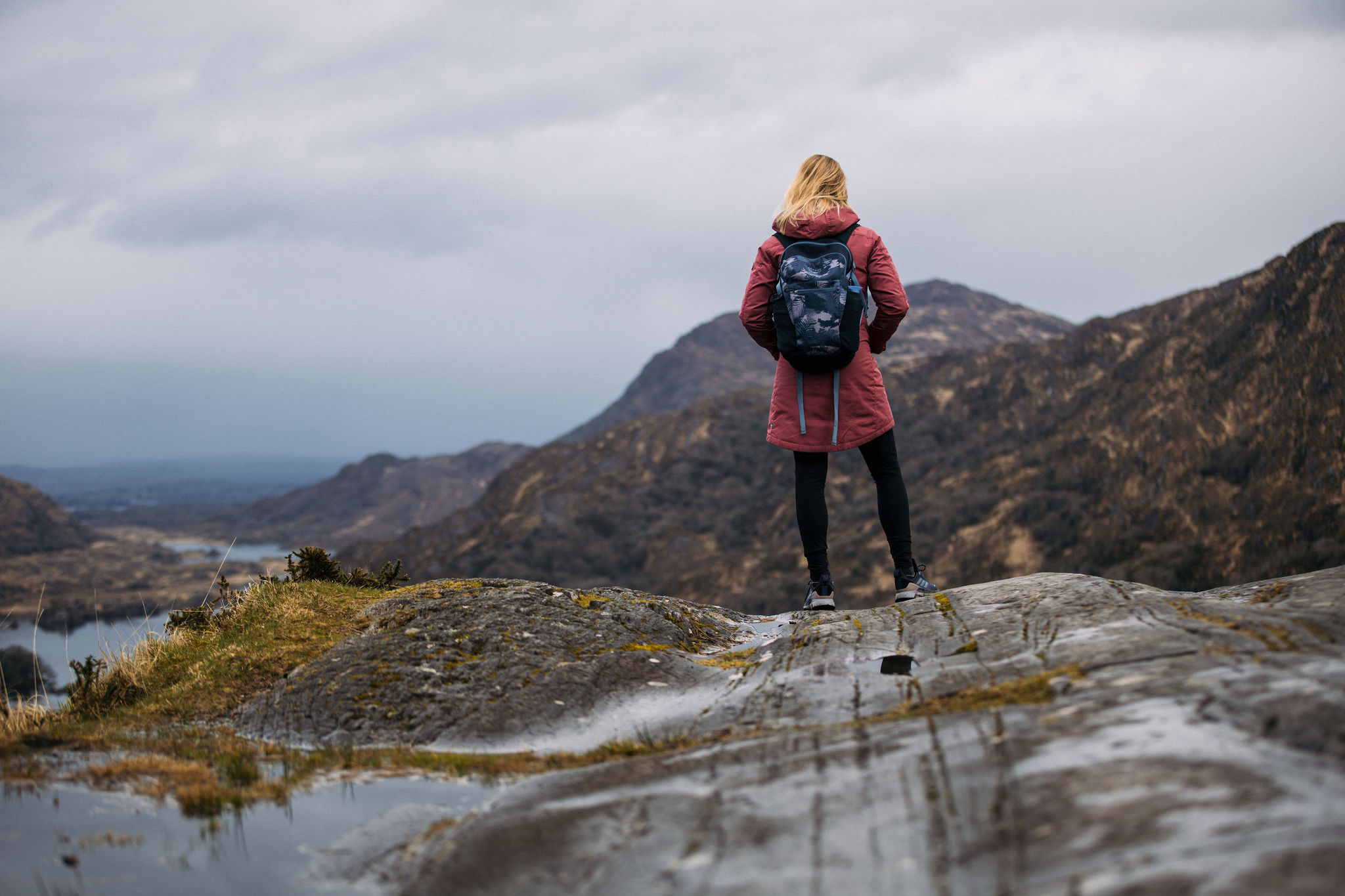 Een persoon in een rood jasje en zwarte leggings staat op een rotsachtige heuveltop tijdens hun rondreis Ierland, uitkijkend over een schilderachtig landschap met verre bergen en een bewolkte lucht. Ze hebben een blauwe rugzak en kijken weg, richting het uitzicht, met gras en een kleine plas op de voorgrond. | Echt Ierland Vakanties