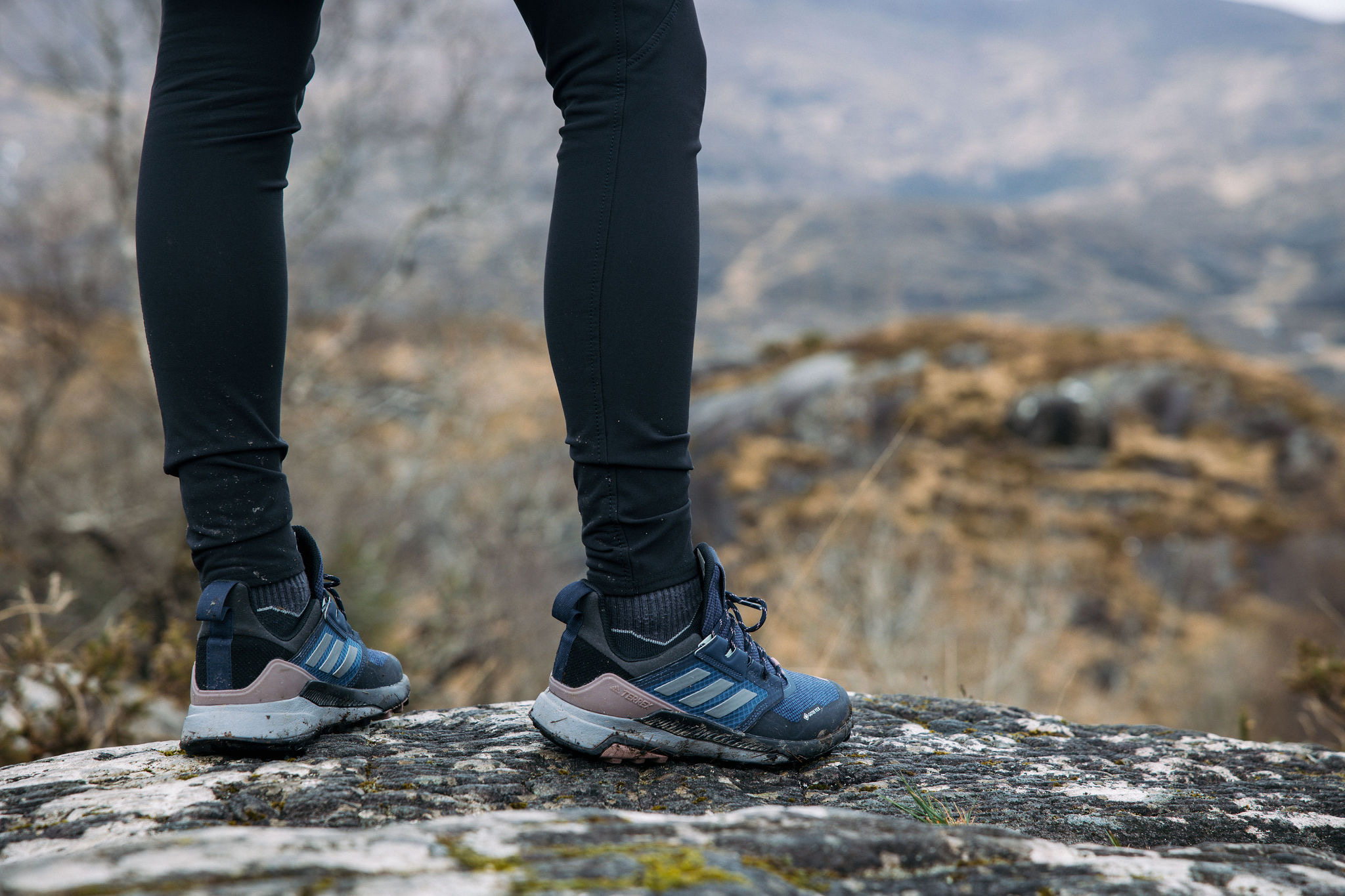 Een persoon in zwarte leggings en blauwe wandelschoenen staat op een rotspunt, uitkijkend over de wazige, aardse heuvels. Deze betoverende scène roept een gevoel van buitenavontuur en verkenning op, vergelijkbaar met een onvergetelijke 'vakantie Ierland'. | Echt Ierland Vakanties