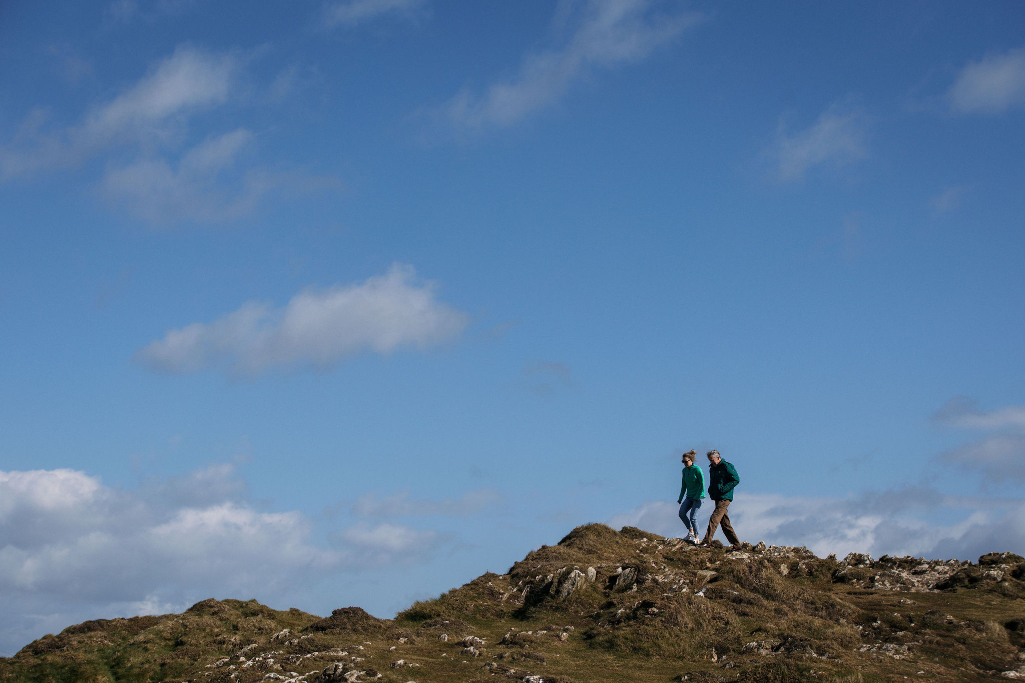 Twee mensen slenteren over een rotsachtige heuveltop onder een helderblauwe lucht met verspreide wolken, en omarmen de ruige schoonheid van Ierland vakanties. Gekleed in casual outdoor kleding en jassen, verkennen ze het grasachtige, steenachtige landschap dat perfect contrasteert met de heldere lucht - een perfecte dag voor avontuur. | Echt Ierland Vakanties