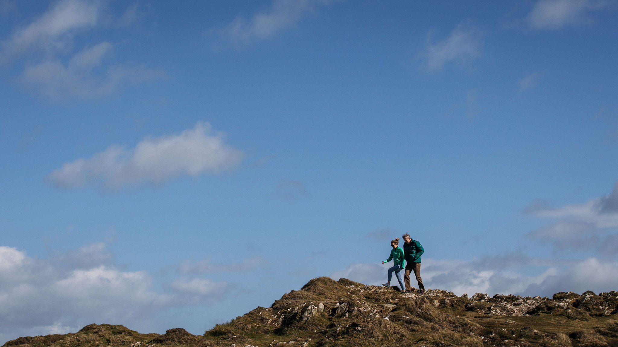 Twee mensen in wandelkleding staan op een rotsachtige heuveltop onder een helderblauwe lucht met verspreide wolken. Ze lijken te genieten van het uitzicht, met ruig terrein dat doet denken aan een rondreis Ierland om hen heen. Het tafereel suggereert een vredig en avontuurlijk moment in de natuur. | Echt Ierland Vakanties