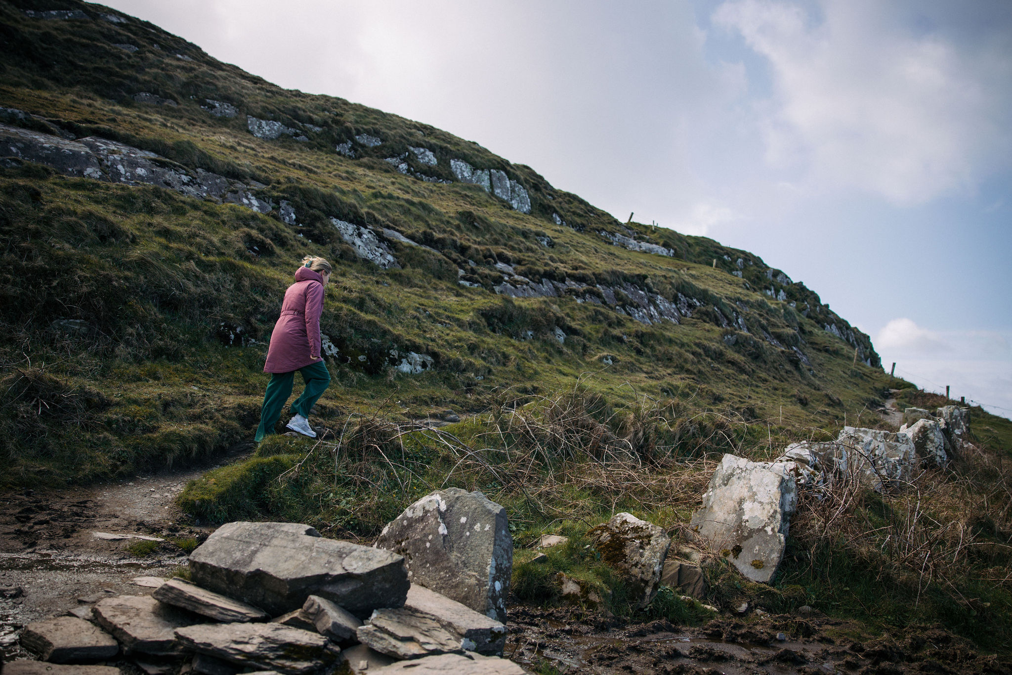 Een persoon in een roze jas en groene broek loopt bergopwaarts op een graspad met rotsen in het ruige Ierland. De helling, bezaaid met stukken gras en stenen onder een bewolkte lucht, straalt rust uit, perfect voor degenen die een outdooravontuur overwegen tijdens hun vakantie in Ierland. | Echt Ierland Vakanties