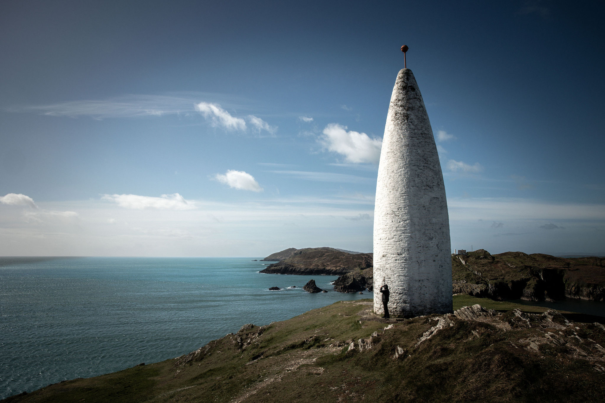 Een witte stenen toren met een kegelvorm staat trots op een klif, uitkijkend over de serene turquoise oceaan - een waar kenmerk van elke rondreis Ierland. De lucht is helder met verspreide wolken en het ruige landschap heeft stukken groen gras. Een eenzame figuur aan de basis geeft een gevoel van schaal. | Echt Ierland Vakanties