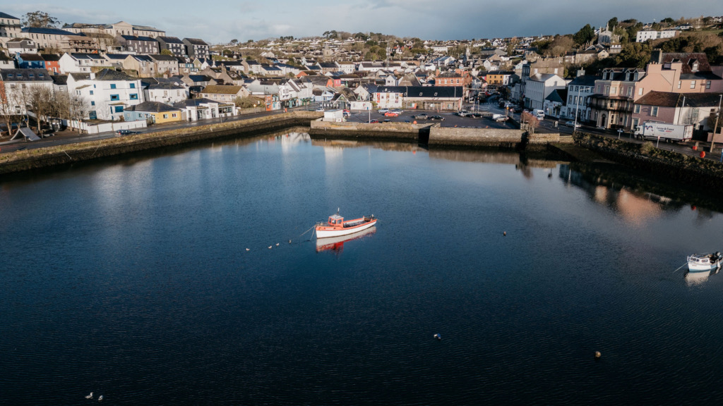Luchtfoto van een serene haven in een schilderachtig Iers stadje, perfect voor degenen die een vakantie naar Ierland overwegen. Een rood-witte boot ligt afgemeerd in het midden, omringd door boeien. De pittoreske haven wordt omlijst door huizen en winkels tegen heuvelachtige landschappen onder een gedeeltelijk bewolkte lucht. | Echt Ierland Vakanties