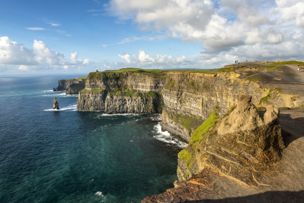 Een schilderachtig uitzicht op de Cliffs of Moher in Ierland, met ruige kliffen die uitkijken over de Atlantische Oceaan. Weelderig groen contrasteert met de diepblauwe zee onder een heldere, deels bewolkte lucht. Mensen dwalen langs de rand en leggen onvergetelijke momenten vast op hun vakantie in Ierland. | Echt Ierland Vakanties