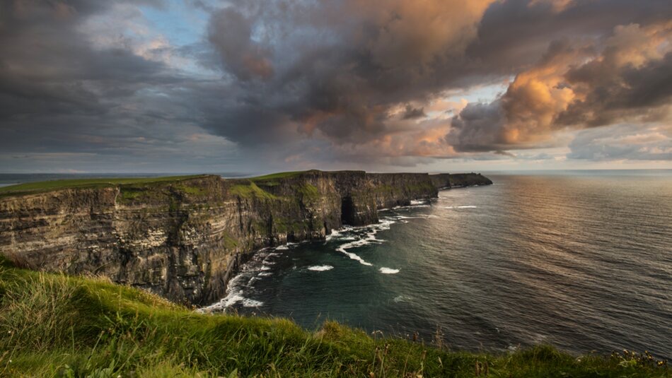 Ervaar de dramatische Cliffs of Moher bij zonsondergang tijdens uw rondreis Ierland. Torenhoog boven de Atlantische Oceaan, golven beuken beneden terwijl donkere wolken gloeien met gouden tinten. Groen gras kroont de kliffen, perfect voor onvergetelijke Ierland vakanties tegen dit ruige landschap. | Echt Ierland Vakanties