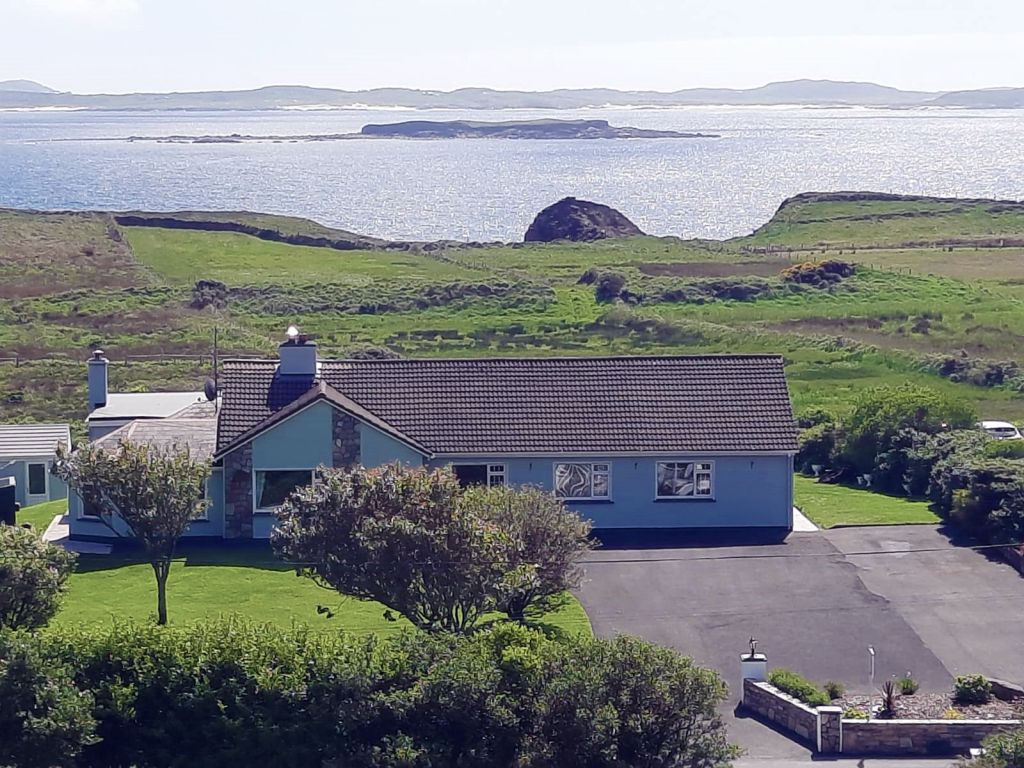 Een blauw, gelijkvloers huis met een grijs dak is omgeven door bomen en een groen gazon. Een geplaveide oprit leidt naar het huis. Op de achtergrond is er een schilderachtig uitzicht op de oceaan en glooiende heuvels onder gedeeltelijk bewolkte luchten - perfect voor degenen die dromen van een vakantie in Ierland. Er is een eiland zichtbaar aan de overkant van het water. | Echt Ierland Vakanties