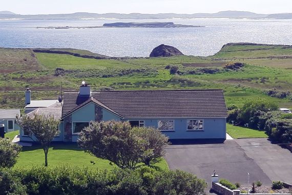 Een blauw, gelijkvloers huis met een grijs dak is omgeven door bomen en een groen gazon. Een geplaveide oprit leidt naar het huis. Op de achtergrond is er een schilderachtig uitzicht op de oceaan en glooiende heuvels onder gedeeltelijk bewolkte luchten - perfect voor degenen die dromen van een vakantie in Ierland. Er is een eiland zichtbaar aan de overkant van het water. | Echt Ierland Vakanties