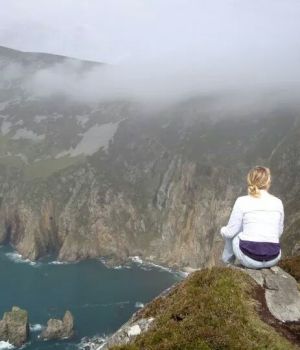 Een persoon met blond haar zit op een met gras begroeide klifrand, uitkijkend over de dramatische kustlijn van Nordirland - perfect voor een Fly & Drive avontuur - met steile kliffen, verpletterende golven en in mist gehulde bergen die een serene, contemplatieve sfeer creëren. | Echt Ierland Vakanties