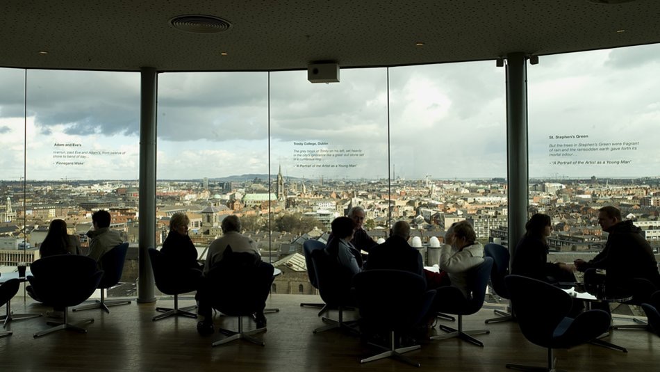 Mensen zitten in silhouet in een ronde lounge vol ramen met uitzicht op een stadsgezicht, wat doet denken aan een stedelijke ontsnapping voordat ze op vakantie gaan naar Ierland. De bewolkte lucht vormt de achtergrond terwijl moderne stoelen een warme, ontspannen sfeer creëren, met tekst in het raam die verschillende uitzichten beschrijft. | Echt Ierland Vakanties