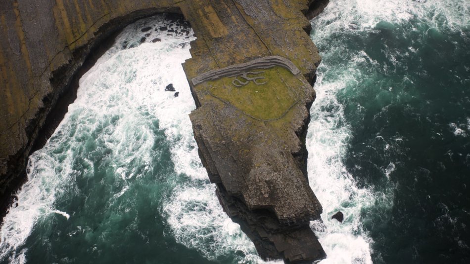 Luchtfoto van een rotsachtige kustpromontory omringd door turbulente oceaan golven, perfect voor een avontuurlijke rondreis Ierland. Groen gras kroont de kliffen, met golfpatronen geëtst in de rotsen. Beneden vermengt diepblauw water zich met schuimend wit, wat de dramatische kustlijn van Ierland laat zien. | Echt Ierland Vakanties