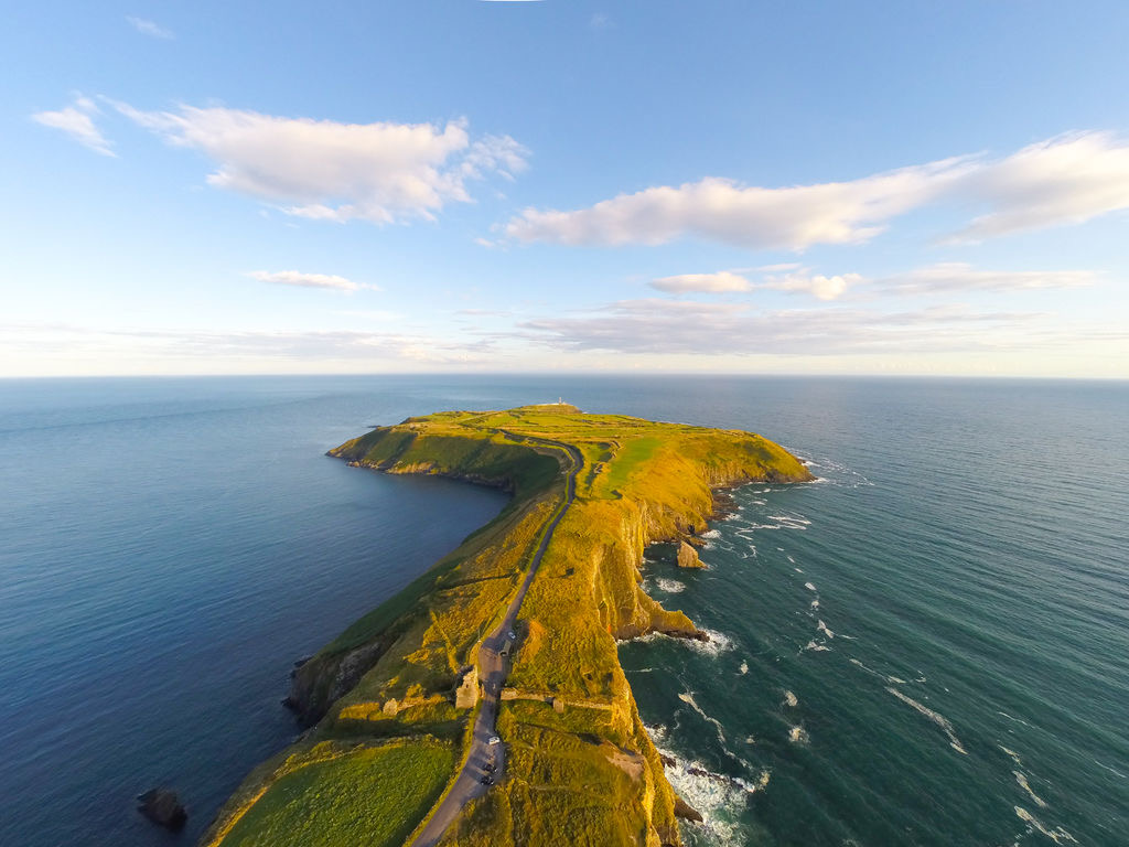 Luchtfoto van een smal, grasachtig schiereiland dat zich uitstrekt in de oceaan, onder een helderblauwe lucht met verspreide wolken. Perfect voor een rondreis Ierland, het kronkelende pad volgt de contouren van het land, geflankeerd door rotsachtige kliffen. Golven klotsen zachtjes tegen de kust, waardoor land en zee contrasteren. | Echt Ierland Vakanties