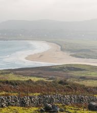 Ervaar het betoverende kustlandschap van Ierland op uw rundreise Irland, met brede zandstranden en zachte golven. Grasvelden en een stenen muur leiden naar verre heuvels en een charmant dorp, alles onder een bewolkte lucht in zacht, natuurlijk licht. | Echt Ierland Vakanties