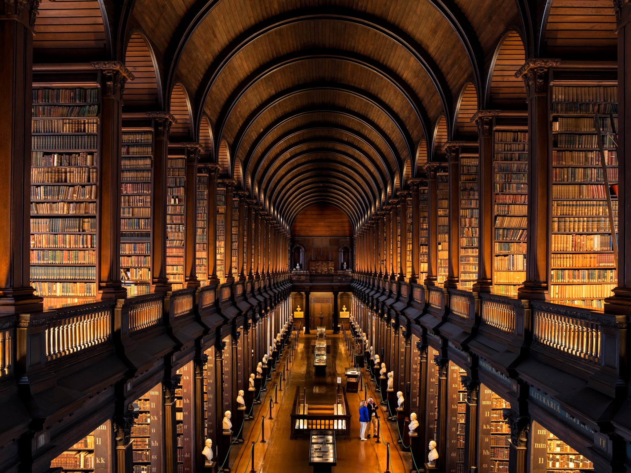De sierlijke bibliotheekhal in Ierland, met zijn gewelfde houten plafond, rijen boeken en bustes langs de zijkanten, biedt een glimp van grandeur en historische elegantie. Warme verlichting verlicht de planken terwijl twee figuren de centrale loopbrug verkennen te midden van glazen vitrines - een perfecte stop tijdens uw vakantie in Ierland. | Echt Ierland Vakanties