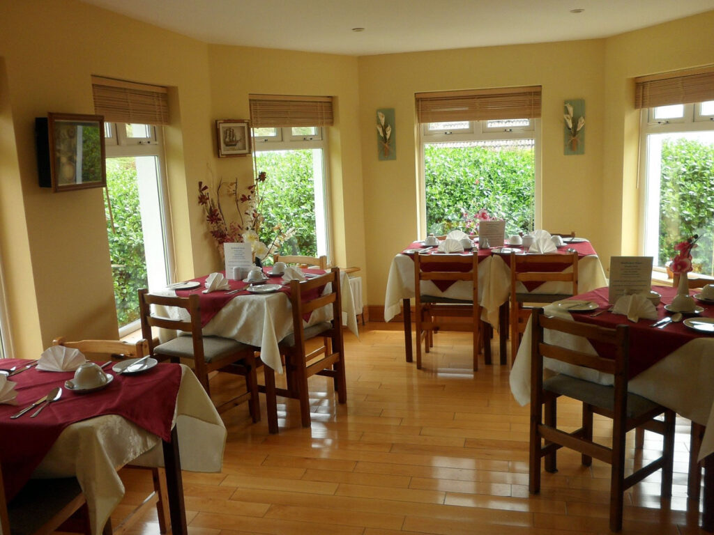 Een gezellige eetkamer die doet denken aan een serene vakantie in Ierland, met houten tafels versierd met witte en bordeauxrode tafellakens. Borden, servetten en kopjes zijn netjes gerangschikt. Grote ramen verwelkomen natuurlijk licht en onthullen weelderig groen buiten tegen de lichte houten vloer. | Echt Ierland Vakanties
