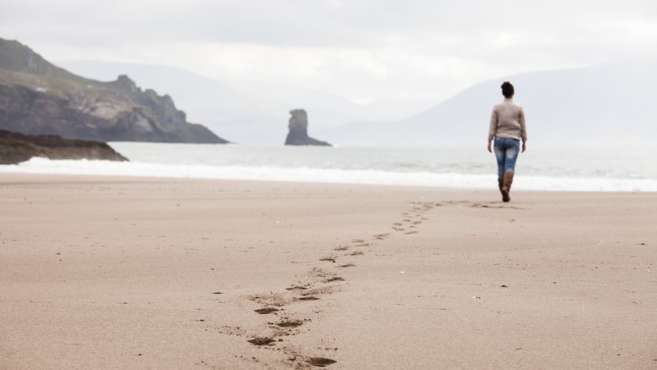 Een persoon wandelt weg op een uitgestrekt, leeg strand in Ierland en laat een spoor van voetafdrukken achter in het natte zand. Ze dragen jeans en een trui. De kalme oceaan ontmoet zachte golven tegen de rotsachtige, bergachtige kustlijn onder een bewolkte lucht - een ideaal tafereel voor een inspirerende vakantie in Ierland. | Echt Ierland Vakanties
