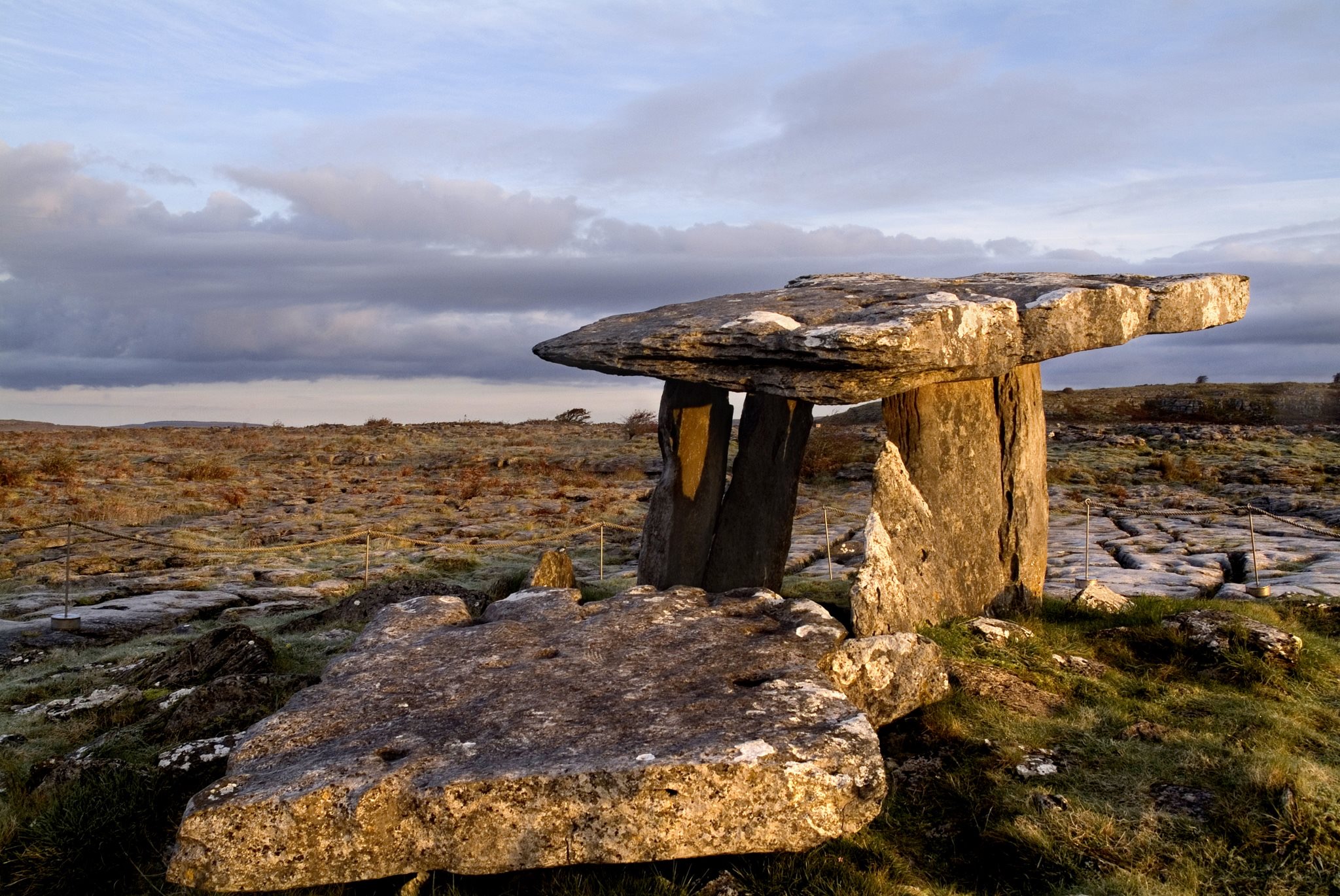 Een verweerde stenen dolmen staat in een grasveld, omgeven door rotsachtig terrein onder een bewolkte lucht - een gezicht dat typerend is voor neolithische architectuur. Perfect voor degenen die een rondreis Ierland plannen, het wilde en ruige landschap komt tot leven met laag zonlicht dat lange schaduwen werpt. | Echt Ierland Vakanties