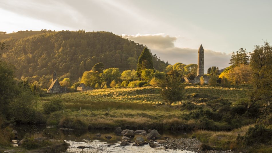 Ontdek een sereen landschap op uw rondreis Ierland, waar een historische site met een ronde toren en oude stenen structuren zich nestelt tussen weelderige bomen. Een rustige rivier stroomt op de voorgrond, rotsen en grasachtige oevers omzomen het pad, terwijl zonlicht een warme gloed werpt over verre heuvels. | Echt Ierland Vakanties