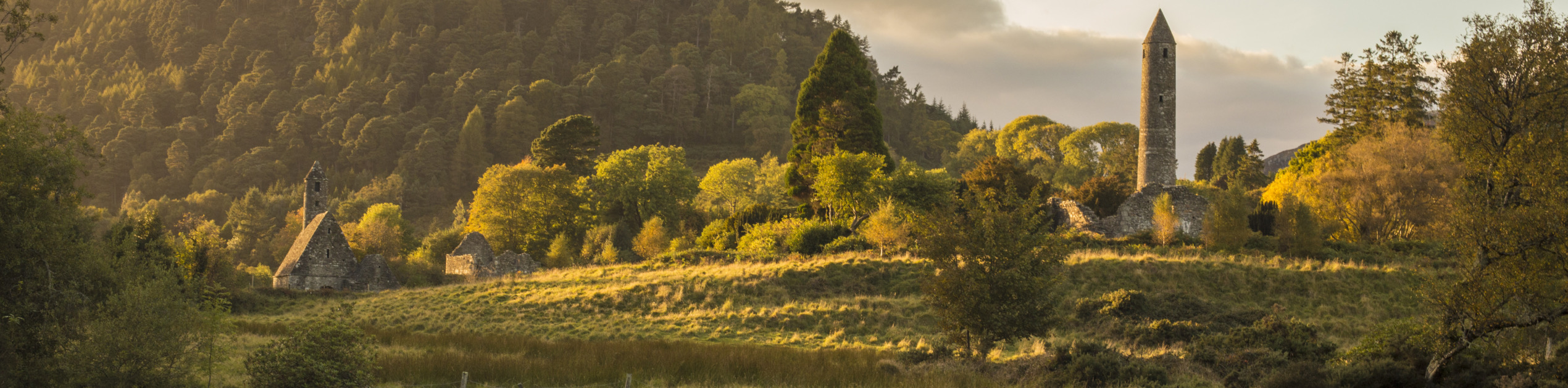 Ontdek een sereen landschap van een weelderige vallei op uw rondreis Ierland. Een stromende beek siert de voorgrond, terwijl oude stenen gebouwen, waaronder een ronde toren en een kerk met een steil dak, zich nestelen tussen de bomen. Het dichte bos op de heuvel baadt in warm zonlicht en legt de aantrekkingskracht van Ierland vakanties vast. | Echt Ierland Vakanties