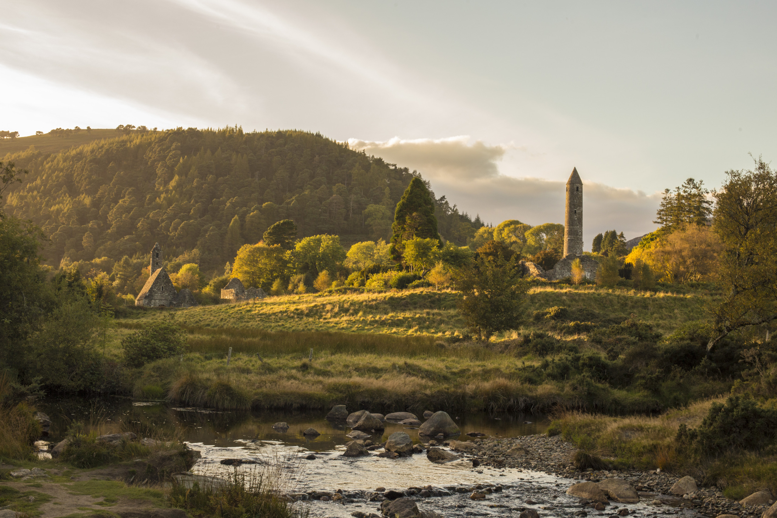 Ontdek een sereen landschap van een weelderige vallei op uw rondreis Ierland. Een stromende beek siert de voorgrond, terwijl oude stenen gebouwen, waaronder een ronde toren en een kerk met een steil dak, zich nestelen tussen de bomen. Het dichte bos op de heuvel baadt in warm zonlicht en legt de aantrekkingskracht van Ierland vakanties vast. | Echt Ierland Vakanties