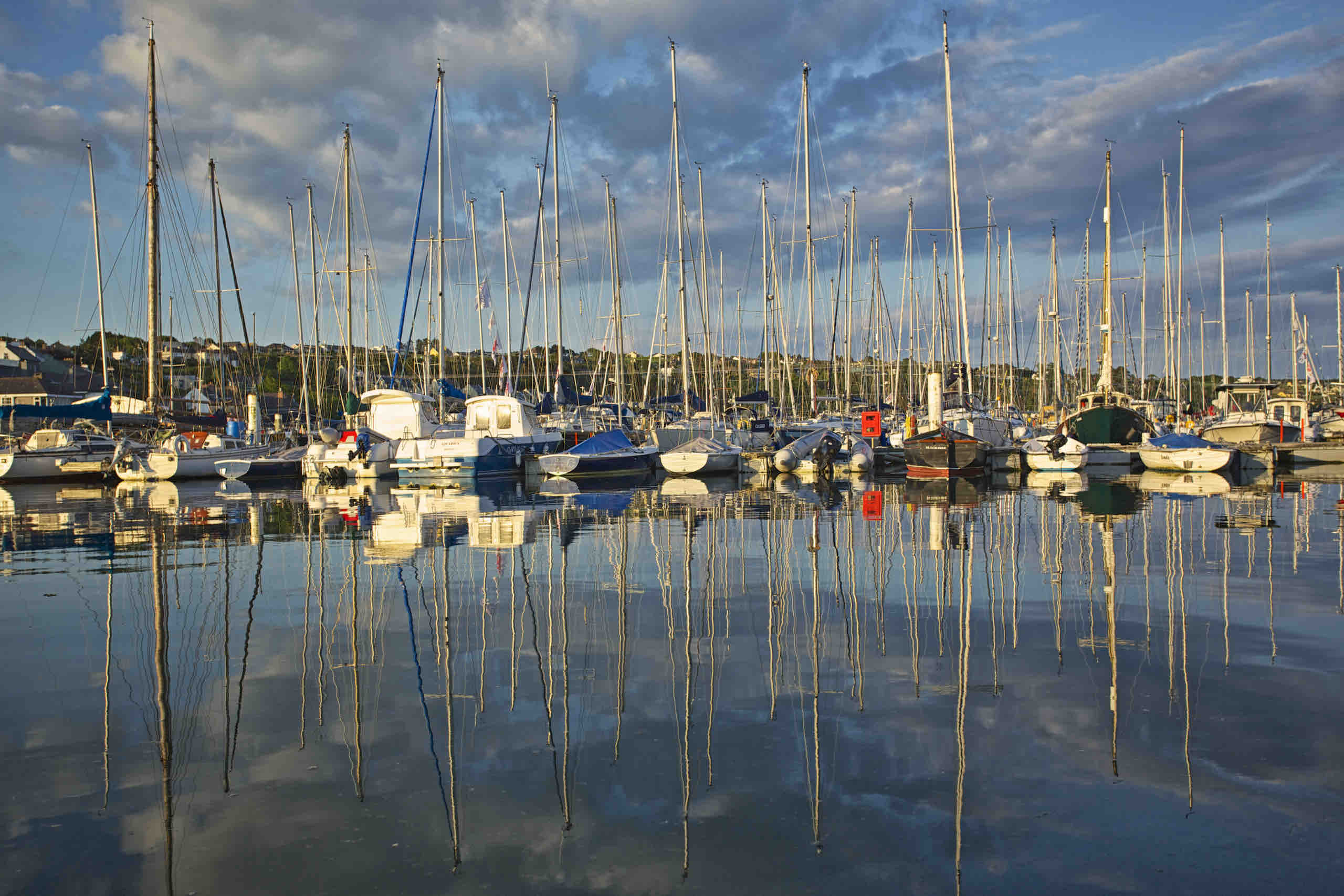 Een kalm havengezicht met talloze zeilboten aangemeerd, hun hoge masten weerspiegelend in het stille water. De lucht is gedeeltelijk bewolkt en de zon werpt een warm licht. Heuvels en gebouwen vormen de achtergrond en versterken de rustige ambiance van deze pittoreske jachthaven, een perfect begin van een vakantieavontuur in Ierland. | Echt Ierland Vakanties