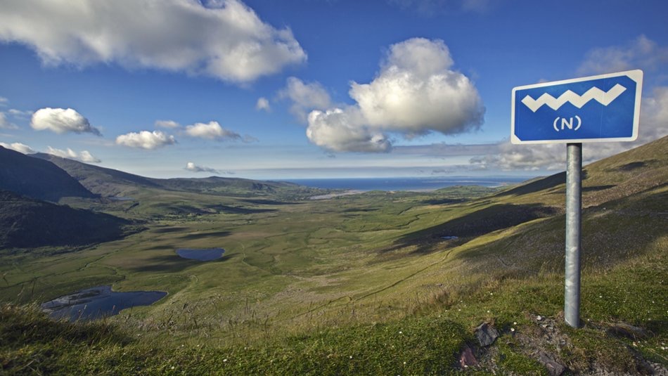 Een schilderachtig landschap ontvouwt zich met een uitgestrekte groene vallei, bezaaid met kleine meren onder een helderblauwe lucht. Op de voorgrond, gemarkeerd door een zigzagwegwijzer met de letter "N", ligt het betoverende pad van uw rondreis Ierland. Heuvels en een verre kustlijn maken dit perfecte vakantie Ierland tafereel compleet. | Echt Ierland Vakanties
