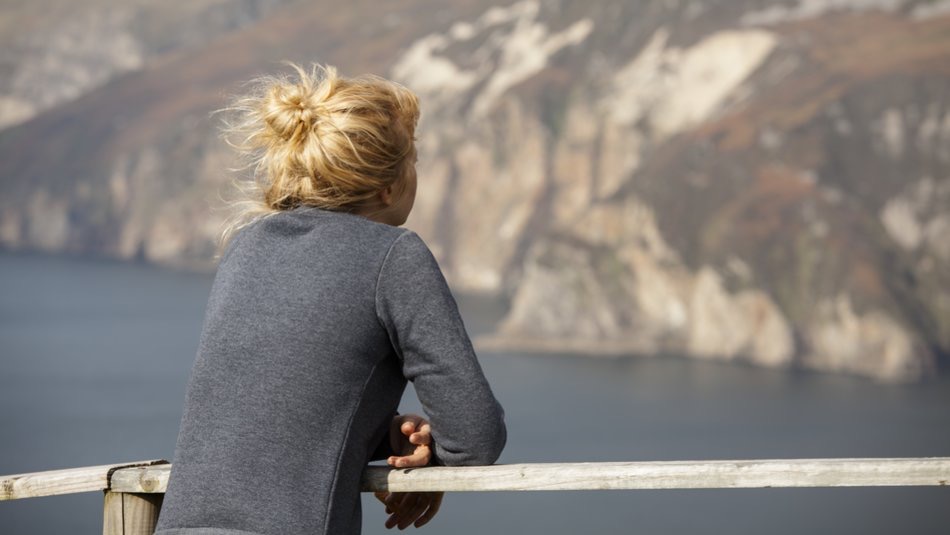 Een persoon met blond haar in een knot leunt op een houten reling en kijkt uit over een schilderachtig uitzicht op de kliffen van Ierland en het kalme water eronder. Ze dragen een grijs shirt met lange mouwen. De kliffen hebben verschillende tinten bruin en grijs, wat een sfeer van serene schoonheid creëert, perfect voor een onvergetelijke vakantie in Ierland. | Echt Ierland Vakanties