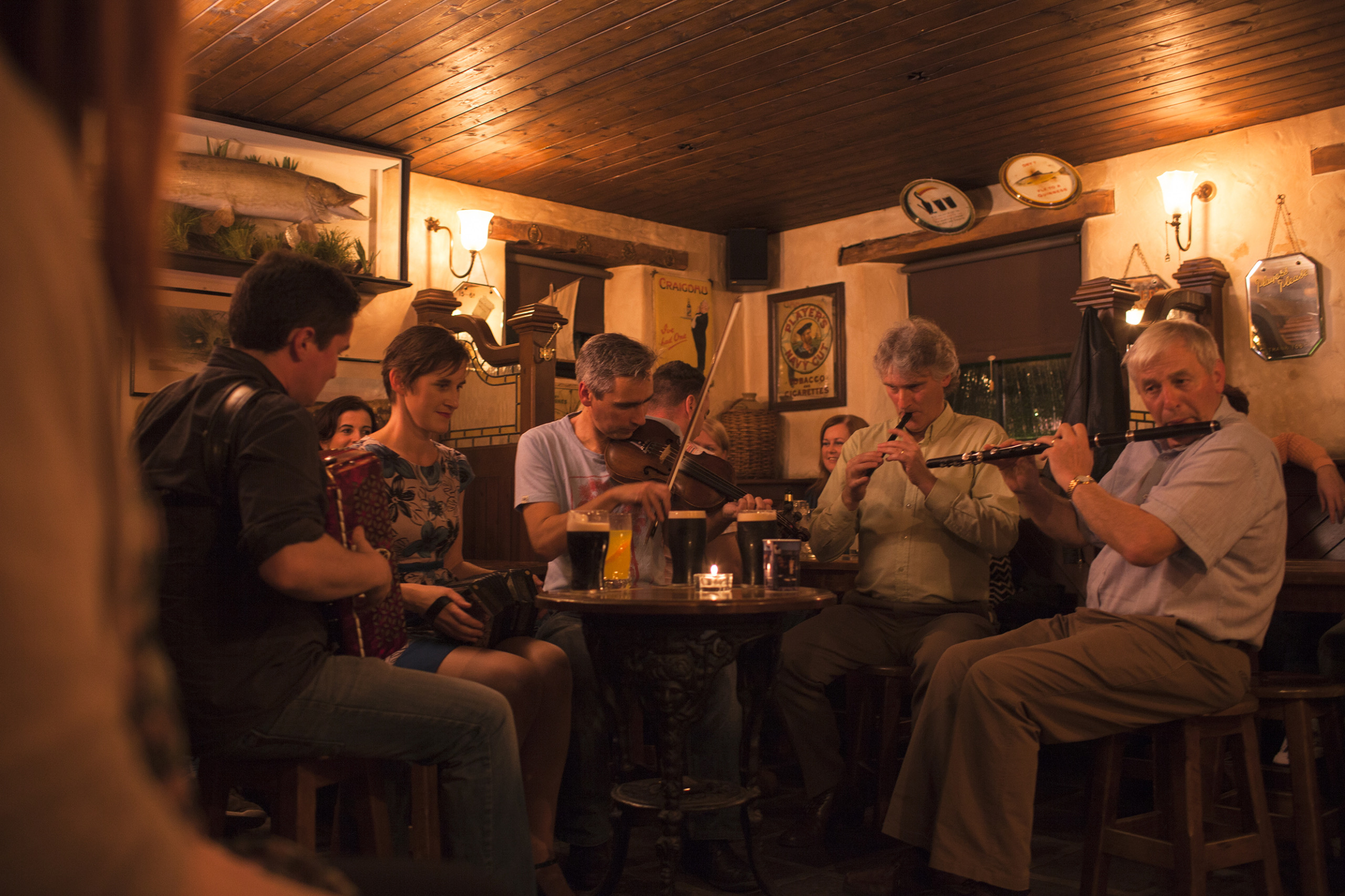 In een gezellige pubscène die doet denken aan onvergetelijke Ierland-vakanties, zitten zes muzikanten in een kring en bespelen traditionele Ierse instrumenten zoals de viool, accordeon en fluiten. De schemerige kamer met houten muren en een grote ingelijste vis boeit gasten die genieten van hun rondreis Ierland-ervaring. | Echt Ierland Vakanties