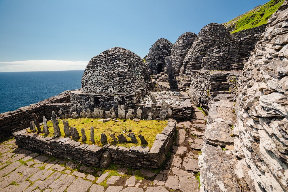 Oude stenen bijenkorfhutten en bouwwerken staan op Skellig Michael, een eiland voor de kust van Ierland, een must-see op elke rondreis door Ierland. Een kleine omheinde begraafplaats met rechtopstaande stenen markeringen is op de voorgrond. De Atlantische Oceaan en een helderblauwe lucht vormen een prachtig decor. | Echt Ierland Vakanties