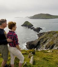 Een stel staat op een met gras begroeide klif in Ierland, met uitzicht op een dramatisch kustlandschap van ruige kliffen en verre groene heuvels. De man draagt een zwart shirt met lange mouwen en een beige broek, terwijl de vrouw een rood geruit shirt en een kaki short draagt. Het is de perfecte scène voor een rondreis Ierland onder kalme zeeën en bewolkte luchten. | Echt Ierland Vakanties