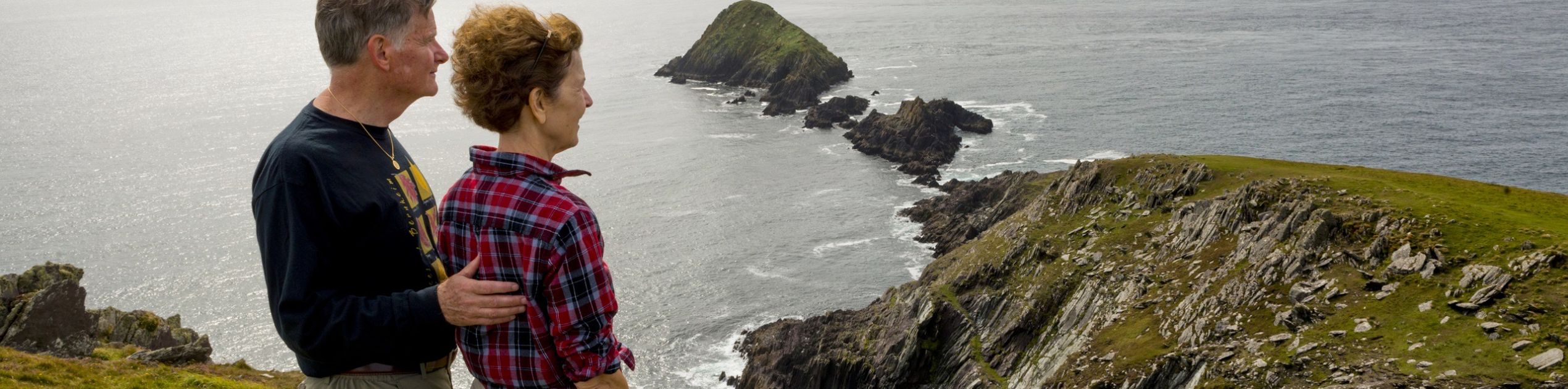 Een stel staat op een met gras begroeide klif in Ierland, met uitzicht op een dramatisch kustlandschap van ruige kliffen en verre groene heuvels. De man draagt een zwart shirt met lange mouwen en een beige broek, terwijl de vrouw een rood geruit shirt en een kaki short draagt. Het is de perfecte scène voor een rondreis Ierland onder kalme zeeën en bewolkte luchten. | Echt Ierland Vakanties