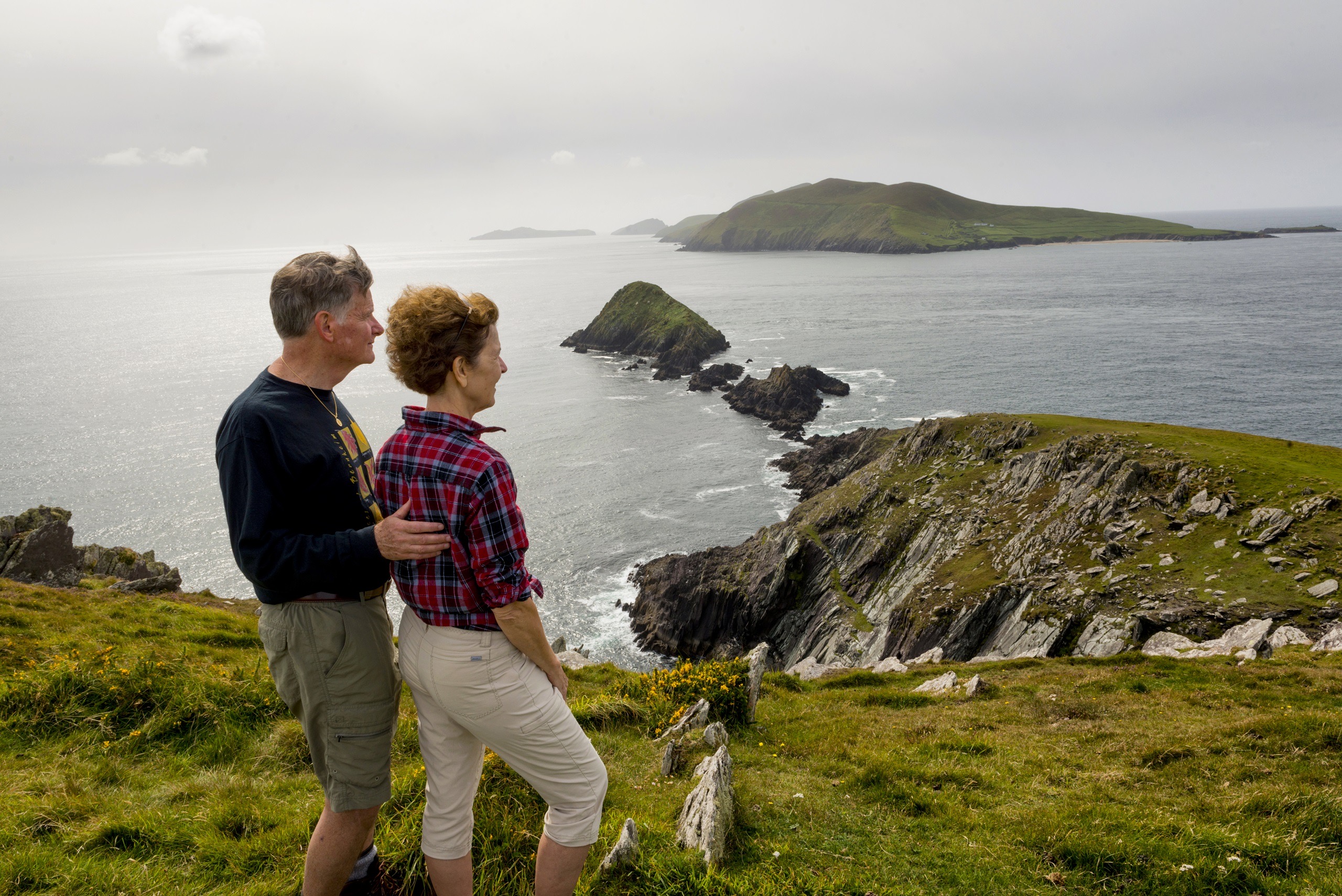 Een stel staat op een met gras begroeide klif in Ierland, met uitzicht op een dramatisch kustlandschap van ruige kliffen en verre groene heuvels. De man draagt een zwart shirt met lange mouwen en een beige broek, terwijl de vrouw een rood geruit shirt en een kaki short draagt. Het is de perfecte scène voor een rondreis Ierland onder kalme zeeën en bewolkte luchten. | Echt Ierland Vakanties