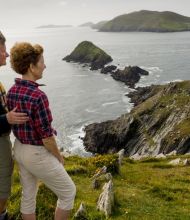 Een stel staat op een met gras begroeide klif, wat de geest van Ierland vakanties belichaamt. De man in een donker shirt raakt lichtjes de rug van de vrouw aan in een rood geruit shirt en beige broek. Samen staren ze naar de rotsachtige kustlijn en het groene eiland onder een bewolkte lucht, terwijl wilde bloemen hun vredige uitzicht bezaaien. | Echt Ierland Vakanties