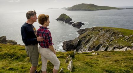 Een stel staat op een met gras begroeide klif, wat de geest van Ierland vakanties belichaamt. De man in een donker shirt raakt lichtjes de rug van de vrouw aan in een rood geruit shirt en beige broek. Samen staren ze naar de rotsachtige kustlijn en het groene eiland onder een bewolkte lucht, terwijl wilde bloemen hun vredige uitzicht bezaaien. | Echt Ierland Vakanties
