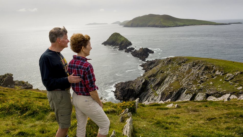 Een stel staat op een met gras begroeide heuvel en geniet van de serene schoonheid van de ruige kustlijn van Ierland tijdens hun vakantie in Ierland. Bewolkte luchten bedekken de kalme oceaan, terwijl rotsachtige kliffen en kleine eilanden dit rustige tafereel compleet maken. Ze delen een rustig moment, vastgelegd in de tijd door hun rondreis Ierland. | Echt Ierland Vakanties