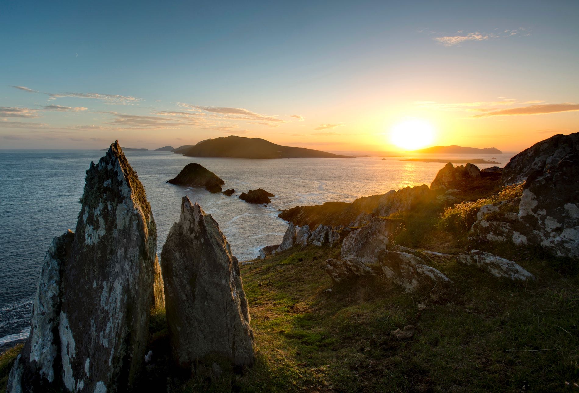 Zonsondergang boven een rotsachtige kustlijn in Ierland met scherpe, grillige kliffen op de voorgrond. De oceaan strekt zich uit tot aan de horizon en weerspiegelt de gouden tinten van de ondergaande zon. Voor degenen die een betoverende rondreis Ierland maken, worden verre eilanden afgetekend tegen een lucht die is bespat met oranje en blauw. | Echt Ierland Vakanties
