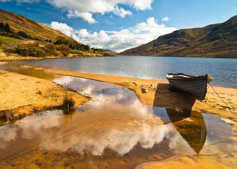 Een sereen landschap dat doet denken aan een idyllische vakantie in Ierland, met een houten boot op een zanderige oever naast een kalm meer. Heuvels omringen het water en weerspiegelen wolken in een blauwe lucht. Zonlicht accentueert het gouden zand en de groene vegetatie op de heuvels, terwijl een smalle stroom het meer in stroomt. | Echt Ierland Vakanties