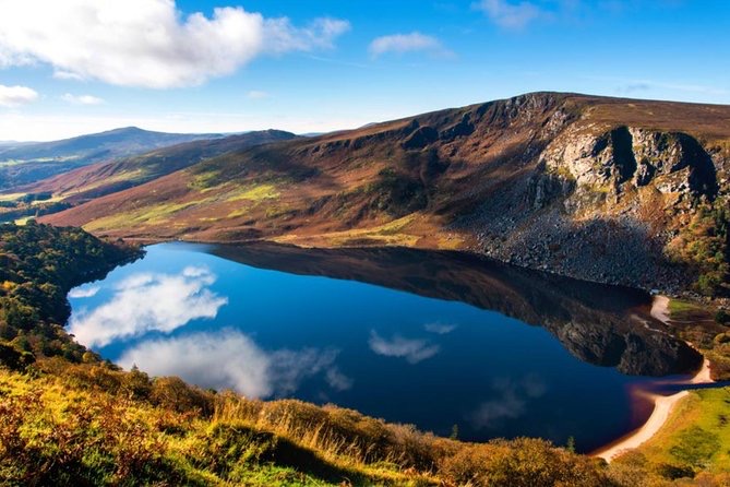 Een sereen meer weerspiegelt de blauwe lucht en verspreide wolken, omringd door ruige heuvels en groen - een perfect tafereel voor een onvergetelijke rondreis Ierland. Verre heuvels koesteren zich in het zonlicht, terwijl weelderig gebladerte de voorgrond siert, met een klein zandgebied dat de rustige wateren van het meer omzoomt. | Echt Ierland Vakanties