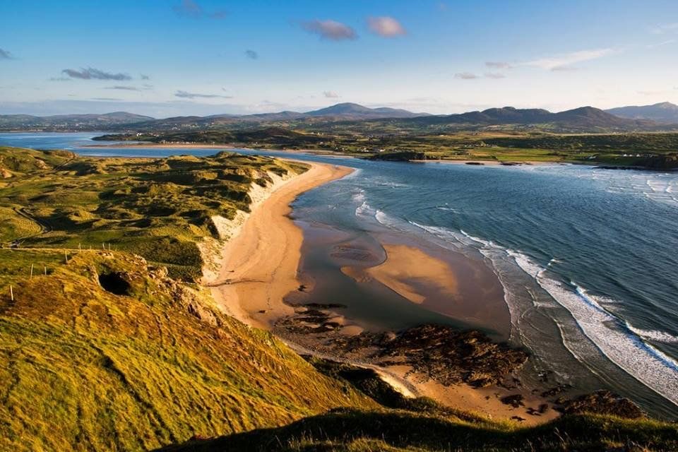 Een prachtig kustlandschap in Ierland heeft een gouden zandstrand dat in de verte kronkelt, perfect voor een vakantie in Ierland. Glooiende groene heuvels omringen de kust, waar zachte oceaan golven het land raken. Bergen rijzen op onder een helderblauwe lucht, bezaaid met donzige wolken. | Echt Ierland Vakanties