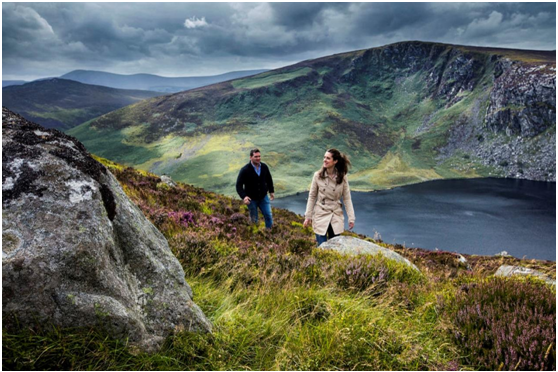 Een man en een vrouw wandelen een met gras begroeide heuvel op in Ierland, omringd door rotsen en heide. Op de achtergrond nestelt een donkerblauw meer zich tussen groene en bruine heuvels onder een bewolkte lucht, wat het avontuur en de natuurlijke schoonheid vastlegt die uniek zijn voor een vakantie in Ierland. | Echt Ierland Vakanties
