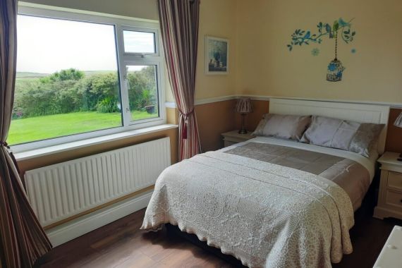 Een gezellige slaapkamer heeft een tweepersoonsbed met een wit dekbed met patroon en bijpassende kussens, die doen denken aan charmante vakanties in Ierland. Beige muren zijn versierd met een muursticker met een vogel en een kooi. Een groot raam met gestreepte gordijnen biedt uitzicht op een weelderige tuin, terwijl de donkere houten vloer de warme tinten van de kamer aanvult. | Echt Ierland Vakanties