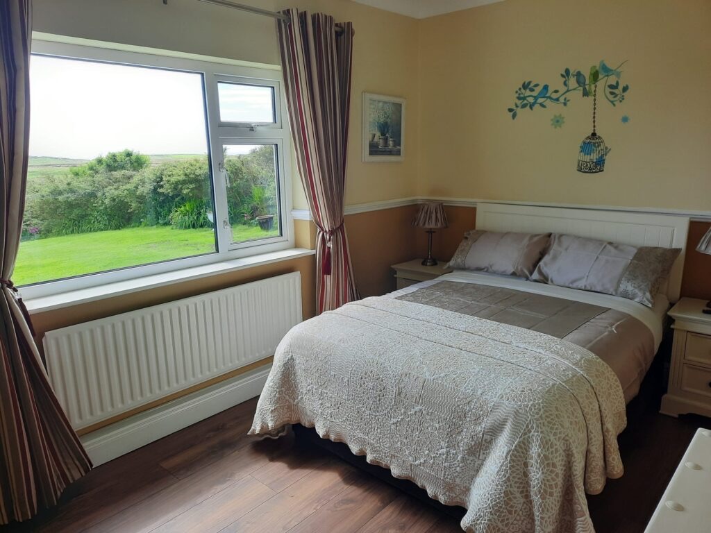 Een gezellige slaapkamer heeft een tweepersoonsbed met een wit dekbed met patroon en bijpassende kussens, die doen denken aan charmante vakanties in Ierland. Beige muren zijn versierd met een muursticker met een vogel en een kooi. Een groot raam met gestreepte gordijnen biedt uitzicht op een weelderige tuin, terwijl de donkere houten vloer de warme tinten van de kamer aanvult. | Echt Ierland Vakanties