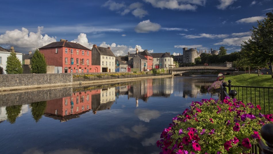 Een rivieroever in Ierland toont kleurrijke gebouwen en weelderig groen dat weerspiegeld wordt in kalm water. Een stenen brug overspant de rivier, met wolken die de blauwe lucht erboven bedekken. Een vakantie in Ierland ervaren betekent dat levendige bloemen contrasteren met dit serene landschap. | Echt Ierland Vakanties