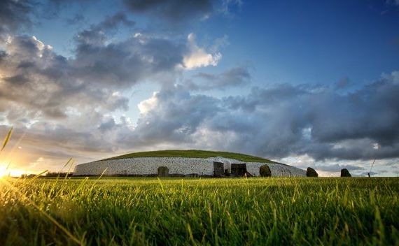 Oude heuvel van Newgrange in Ierland onder een dramatische hemel. De grote, met gras bedekte koepel heeft een witte stenen façade en een ingang. Het zonlicht gluurt over de horizon en verlicht het groene veld - een must-see op elke vakantie in Ierland. Grijze en witte wolken bedekken de blauwe lucht. | Echt Ierland Vakanties