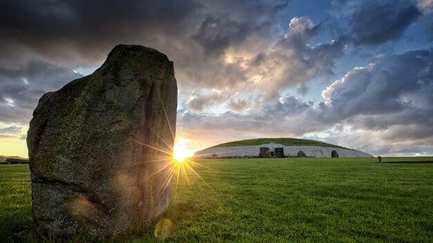 Een stenen monument staat majestueus met een grote staande steen op de voorgrond en een met gras begroeide heuvel op de achtergrond, onder een dramatische zonsondergang. Deze boeiende scène, perfect voor elke vakantie in Ierland, hint naar een oude betekenis, vooral omdat lichtstralen een intrigerende ingang verlichten. | Echt Ierland Vakanties