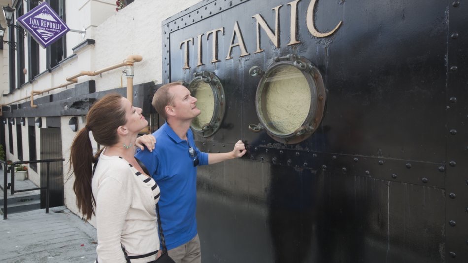 Een man en een vrouw turen door patrijspoorten op een zwarte muur met het opschrift "TITANIC", wat een nostalgische charme oproept die lijkt op een rondreis door Ierland. De man draagt een blauw shirt met een zonnebril om zijn nek, terwijl de vrouw, in haar witte top en paardenstaart, de nonchalante elegantie van vakantie Ierland belichaamt. | Echt Ierland Vakanties