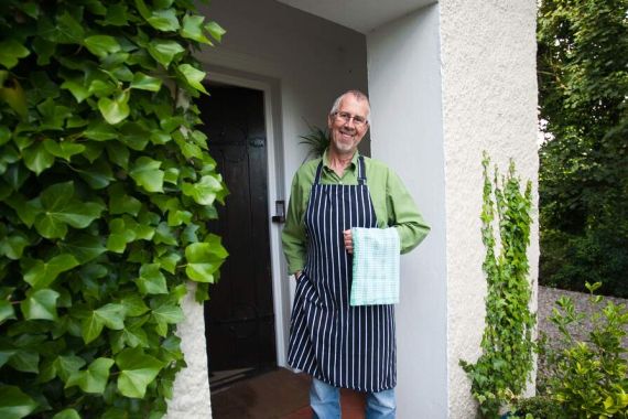 Een glimlachende man staat bij een witte deuropening met een structuur, omlijst door weelderige groene klimop, wat de charme van Urlaub Irland oproept. Hij draagt een bril, een groen shirt, een zwart-wit gestreepte schort, een blauwe spijkerbroek en witte schoenen, en houdt een geblokte handdoek vast. Potplanten omringen de ingang, wat bijdraagt aan de groene sfeer. | Echt Ierland Vakanties