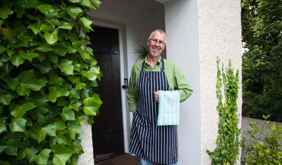 Een glimlachende man staat bij een witte deuropening met een structuur, omlijst door weelderige groene klimop, wat de charme van Urlaub Irland oproept. Hij draagt een bril, een groen shirt, een zwart-wit gestreepte schort, een blauwe spijkerbroek en witte schoenen, en houdt een geblokte handdoek vast. Potplanten omringen de ingang, wat bijdraagt aan de groene sfeer. | Echt Ierland Vakanties
