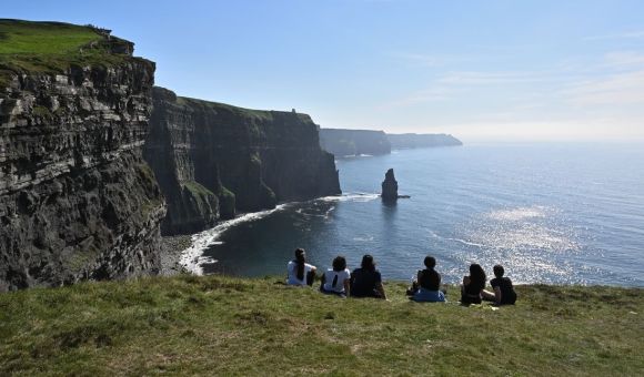 Een groep mensen zit op de rand van een met gras begroeide klif, met uitzicht op de Cliffs of Moher in Ierland. De torenhoge kliffen rijzen steil op uit de Atlantische Oceaan, met kalme golven eronder. Genietend van hun rondreis Ierland, verwonderen ze zich over het pittoreske landschap onder een helderblauwe lucht. | Echt Ierland Vakanties