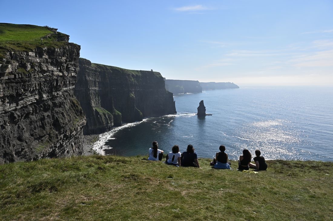Een groep mensen zit op de rand van een met gras begroeide klif, met uitzicht op de Cliffs of Moher in Ierland. De torenhoge kliffen rijzen steil op uit de Atlantische Oceaan, met kalme golven eronder. Genietend van hun rondreis Ierland, verwonderen ze zich over het pittoreske landschap onder een helderblauwe lucht. | Echt Ierland Vakanties