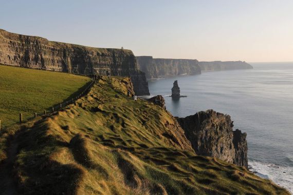 De afbeelding legt de Cliffs of Moher vast tijdens een rundreise in Ierland, tegen een heldere hemel. De dramatische kliffen rijzen majestueus op uit de Atlantische Oceaan, bedekt met weelderig groen gras. Een ruige kustlijn strekt zich ver uit, met een eenzame zeestapel prominent in het midden. | Echt Ierland Vakanties